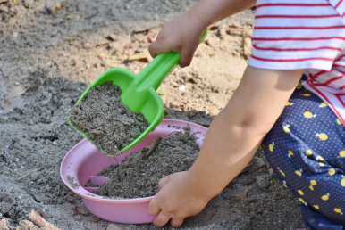 Na obrazku widzimy dłonie dziecka, które nabiera na zieloną szufelkę piasek i wrzuca go do różowej miseczki.